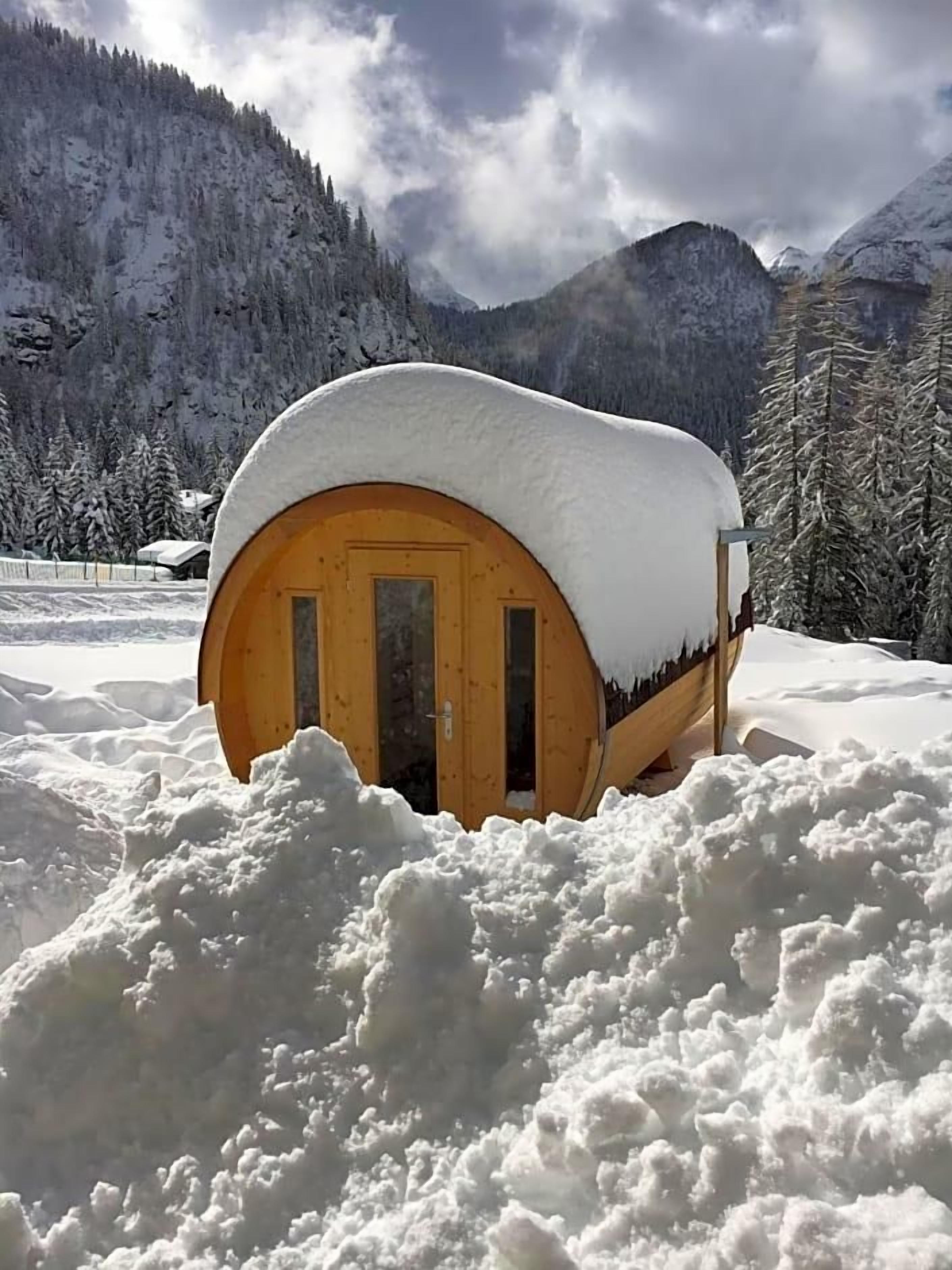 A snow-covered wooden cabin with a door, surrounded by a snowy landscape and mountains.