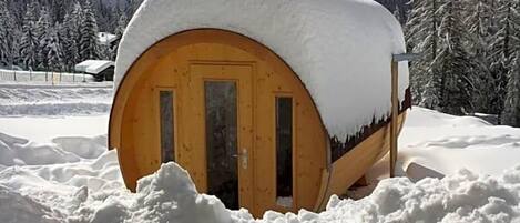 A snow-covered wooden cabin with a door, surrounded by a snowy landscape and mountains.
