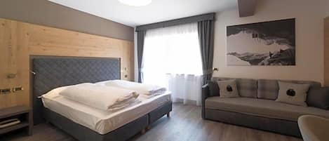 Un dormitorio con cama, sofá, cabecero de madera, un cuadro y una ventana con cortinas.