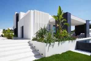 En moderne bygning med hvid facade, swimmingpool og en velholdt have med forskellige planter.