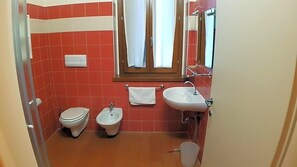 A bathroom with red tiles, a toilet, a bidet, a sink, and a window with curtains. - Ostello Molino Di Basso (Torre Pallavicina)