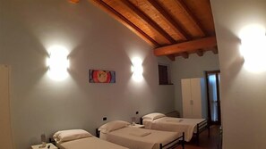 A room with three beds, wooden ceiling beams, a wall-mounted light fixture, and a door leading to another room. - Ostello Molino Di Basso (Torre Pallavicina)