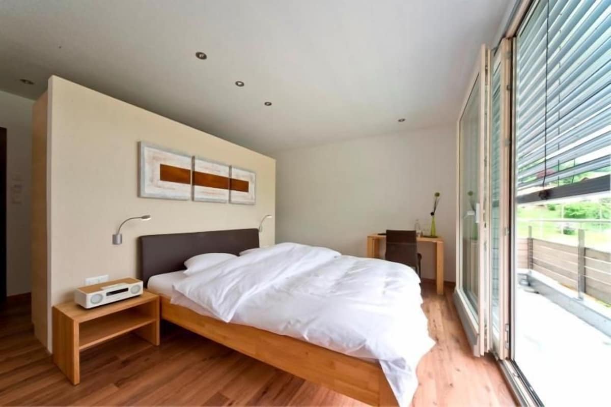 A modern bedroom with a large bed, bedside tables, a wall-mounted artwork, and a sliding glass door leading to an outdoor area.
