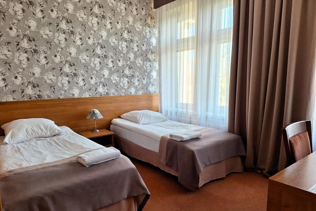 Quarto de hotel com duas camas, uma cabeceira de madeira, mesa de cabeceira com luminária e vista para o exterior através de cortinas finas.