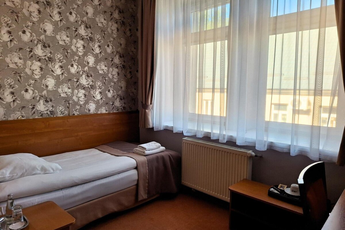 Habitación de hotel con una cama individual, una mesita de noche con lámpara, un radiador y una ventana con cortinas traslúcidas.