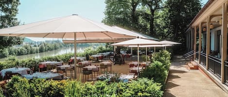 An outdoor dining area with tables and umbrellas, surrounded by greenery and overlooking a body of water.