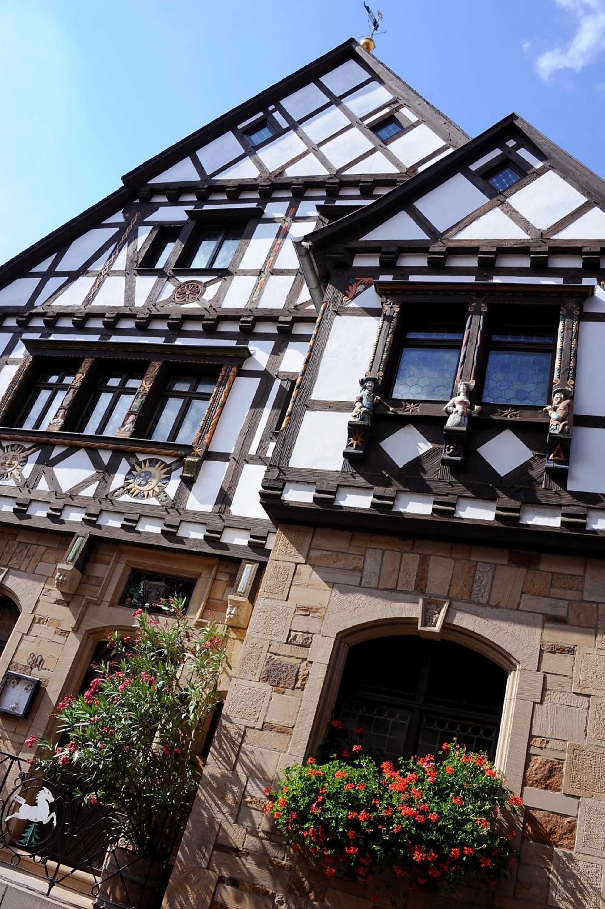 A half-timbered building with a stone base, multiple windows, and a small balcony with statues.