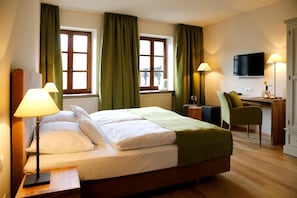 A hotel room with a bed, two windows with curtains, a desk with a chair, a television, and two lamps. - Weinkastell Zum Weissen Ross (Kallstadt)