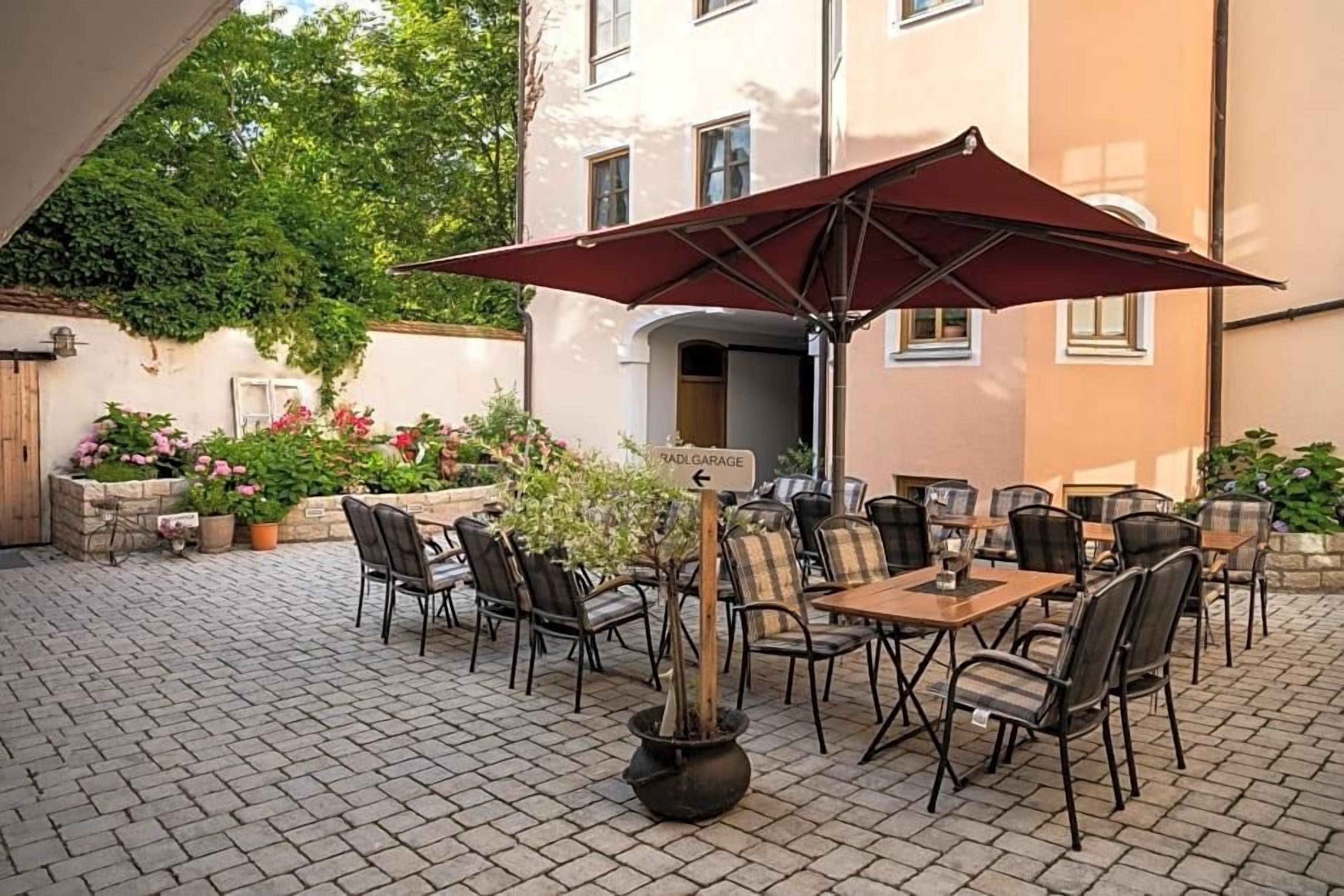 An outdoor patio with tables and chairs, surrounded by potted plants and a building in the background.