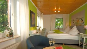 A hotel room with a bed, a chair, a table with a watermelon and wine glasses, and a window with curtains. - Landhaus Heidekrug (Diekholzen)