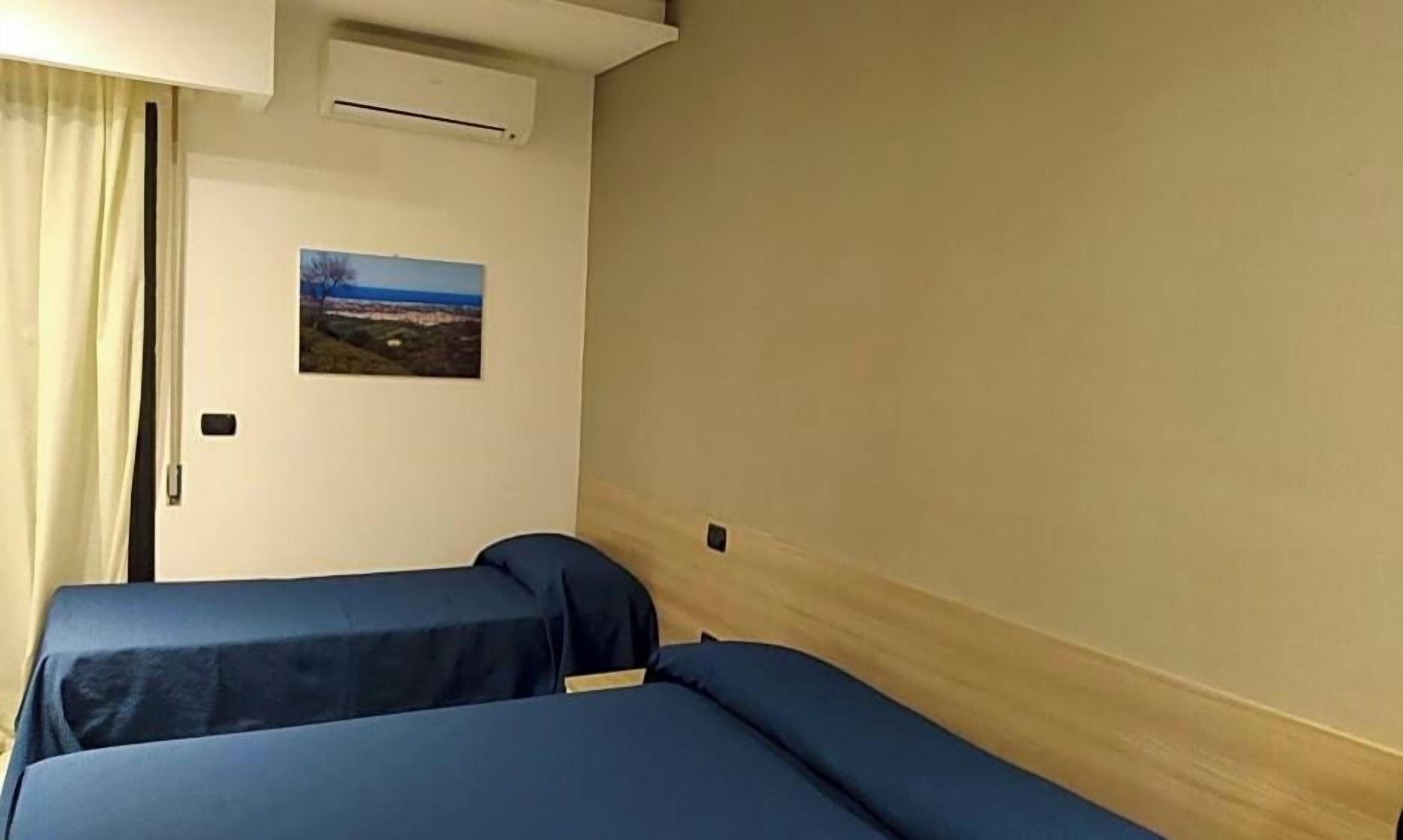 Habitación de hotel con dos camas, una ventana con cortinas, un equipo de aire acondicionado y un cuadro colgado en la pared.