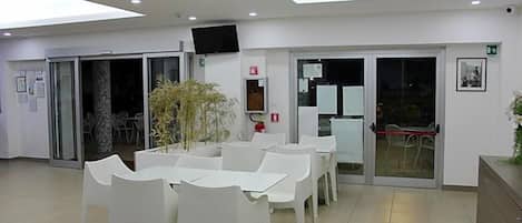 Un espacio interior moderno con zona de comedor con mesas y sillas blancas, un televisor de pantalla plana en la pared y un puerta de cristal que da al exterior.