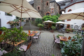Een terras met tafels en stoelen, omgeven door stenen gebouwen en groen.