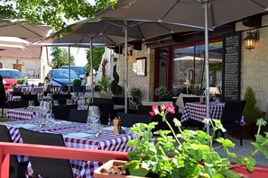 Un'area pranzo all'aperto con tavoli apparecchiati per due, riparata da ombrelloni e una bacheca con il menu a destra.