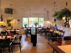 A restaurant interior with wooden flooring, tables with red napkins, hanging yellow pendant lights, and a large black column. - Bredeneeker Gasthaus (Rastorf)