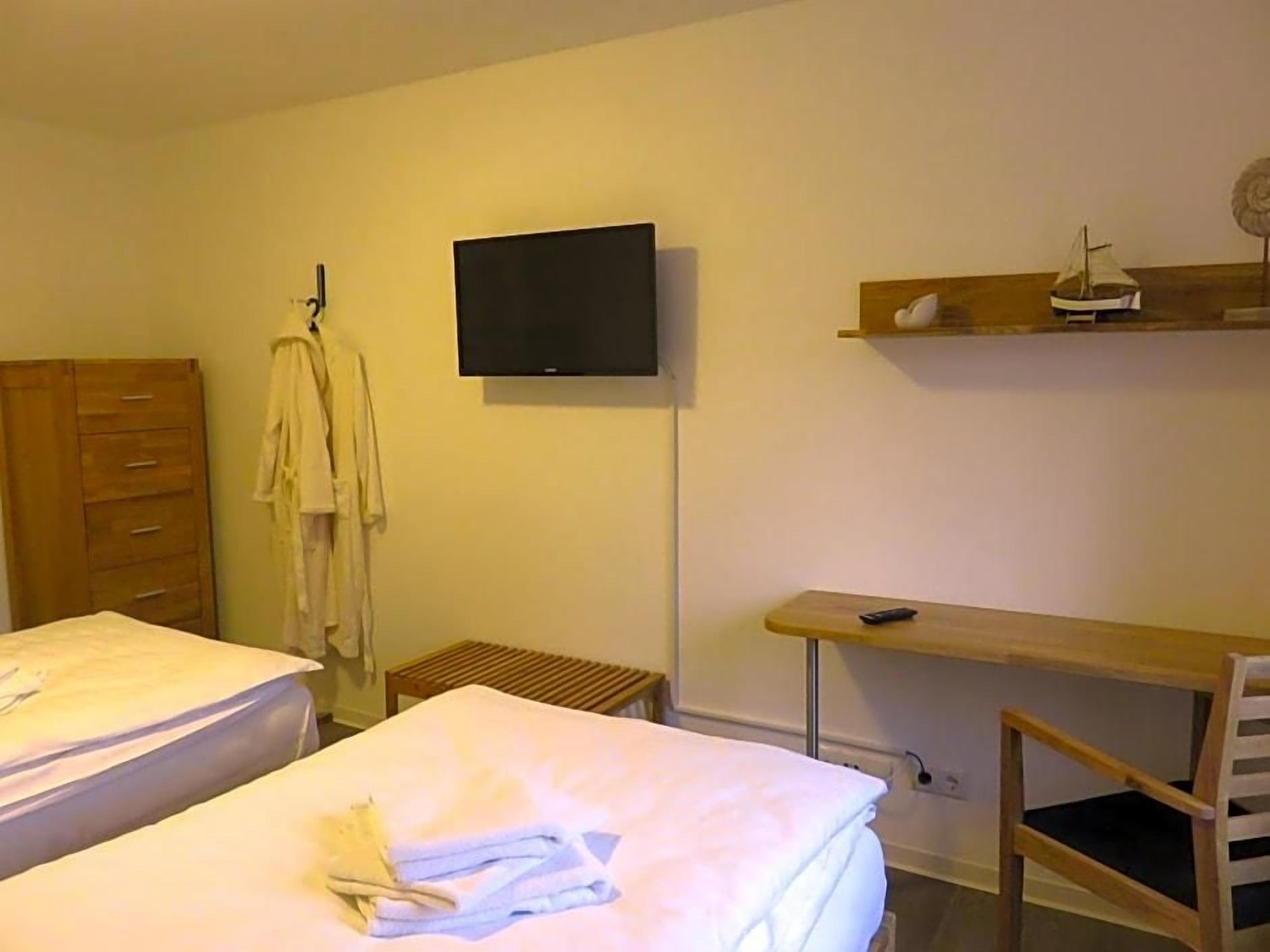 Quarto de hotel com duas camas, televisão fixada na parede, cômoda de madeira e uma pequena mesa com uma cadeira.