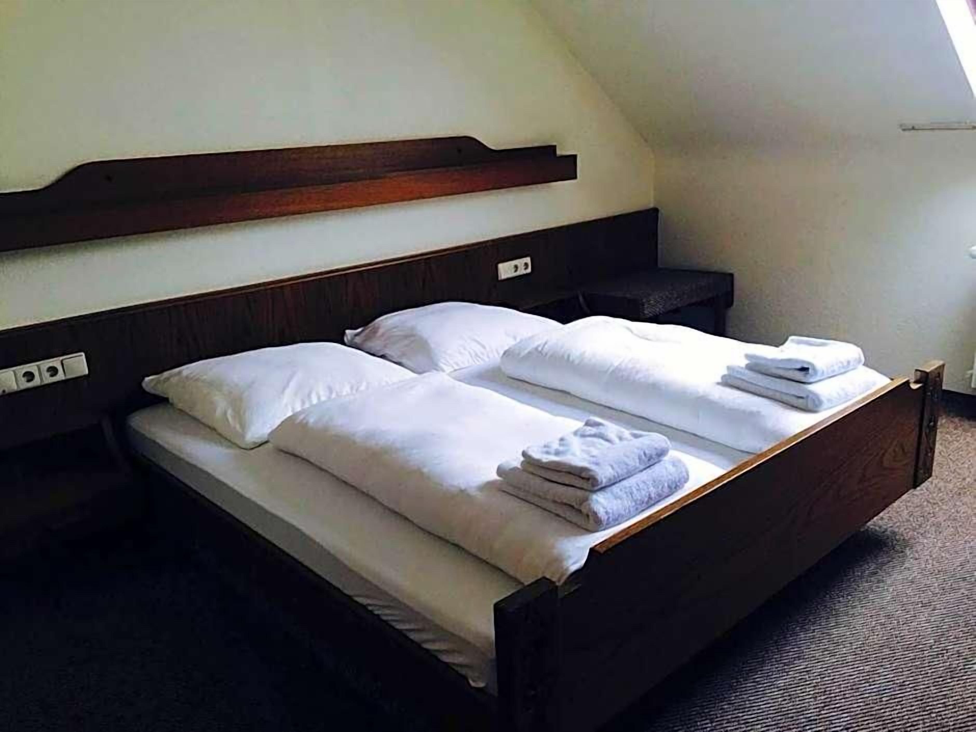 Een tweepersoonsbed met een houten hoofdbord, witte lakens en een opgevouwen handdoek op de zijkant.
