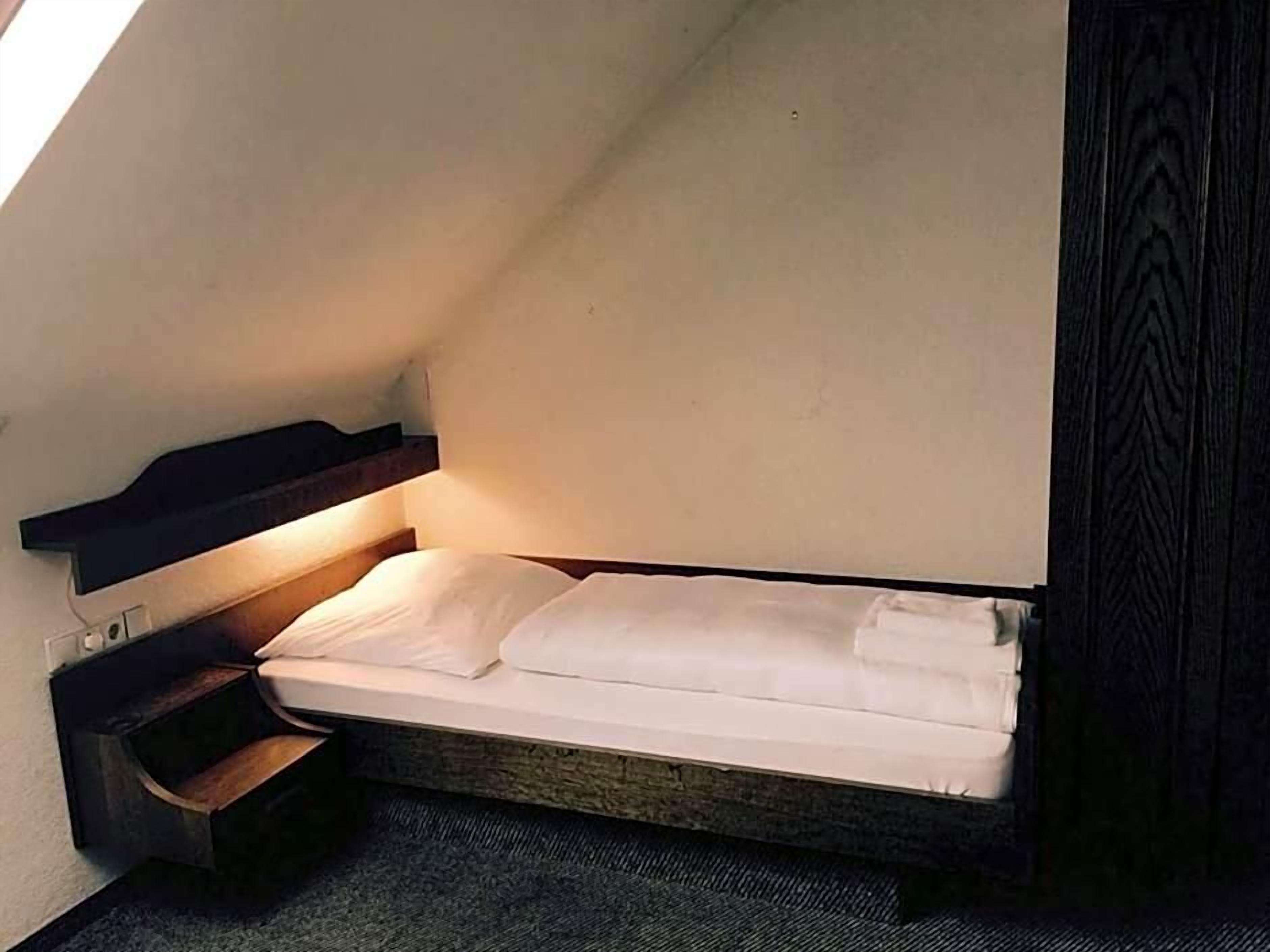 Ein kleiner, schmaler Raum mit einem Bett, einem Nachttisch und einem Wandregal.
