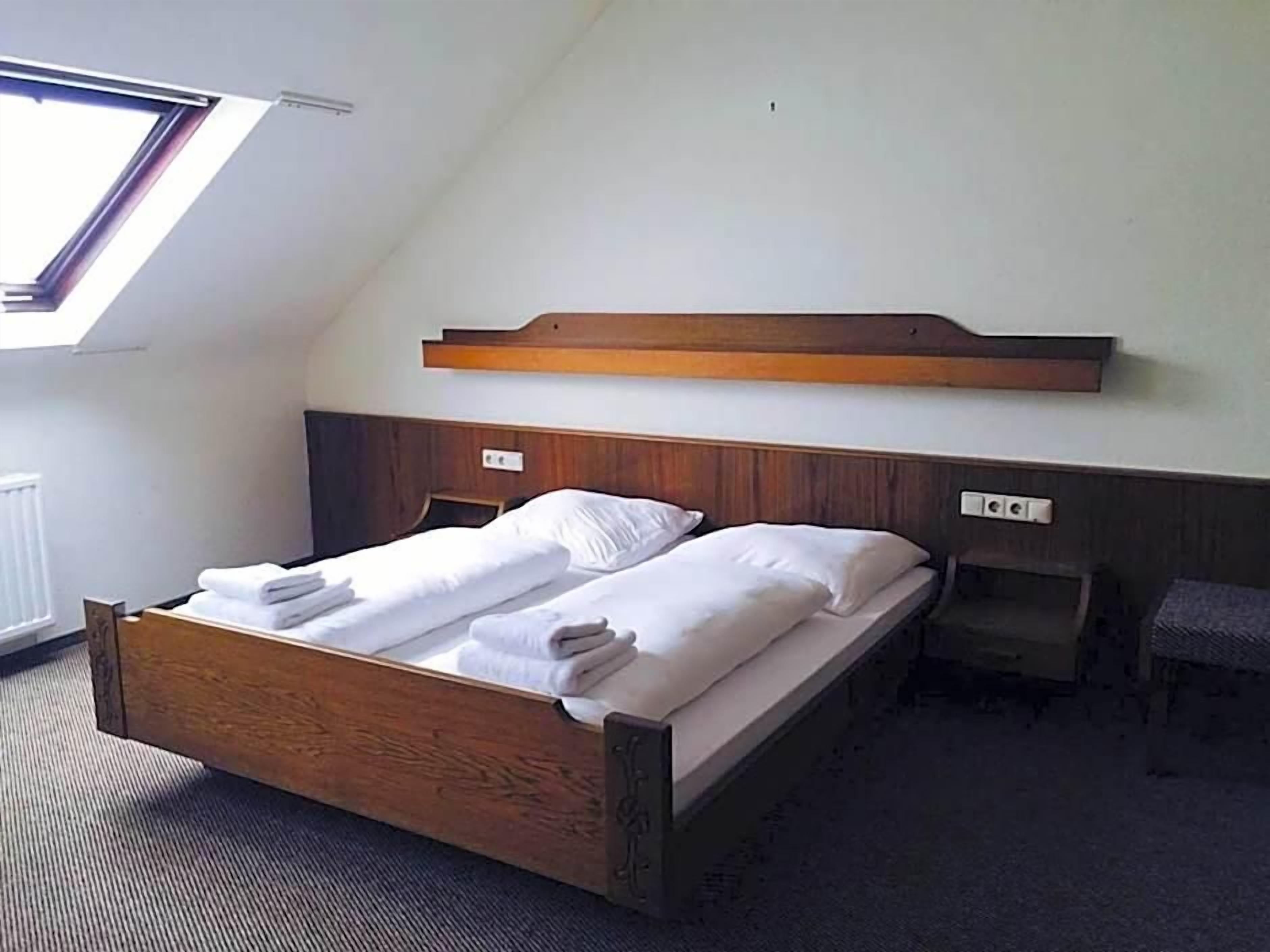 Een enkel bed met houten frame en hoofdbord, een nachtkastje en een kleine stoel in een kamer met een schuine plafond en dakraam.