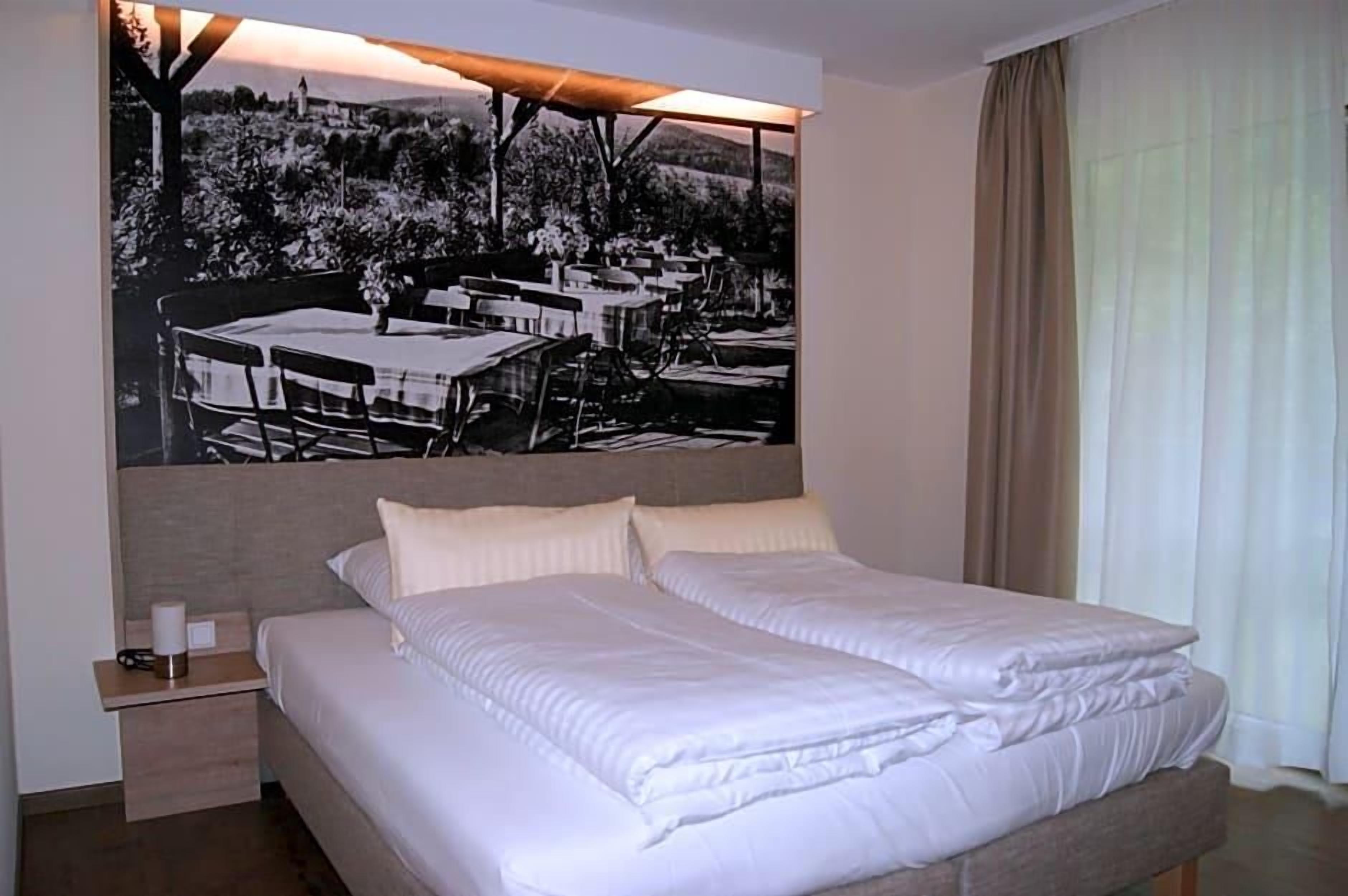 Habitación de hotel con una cama grande, una mesita de noche, una obra de arte en la pared y una ventana con cortinas.