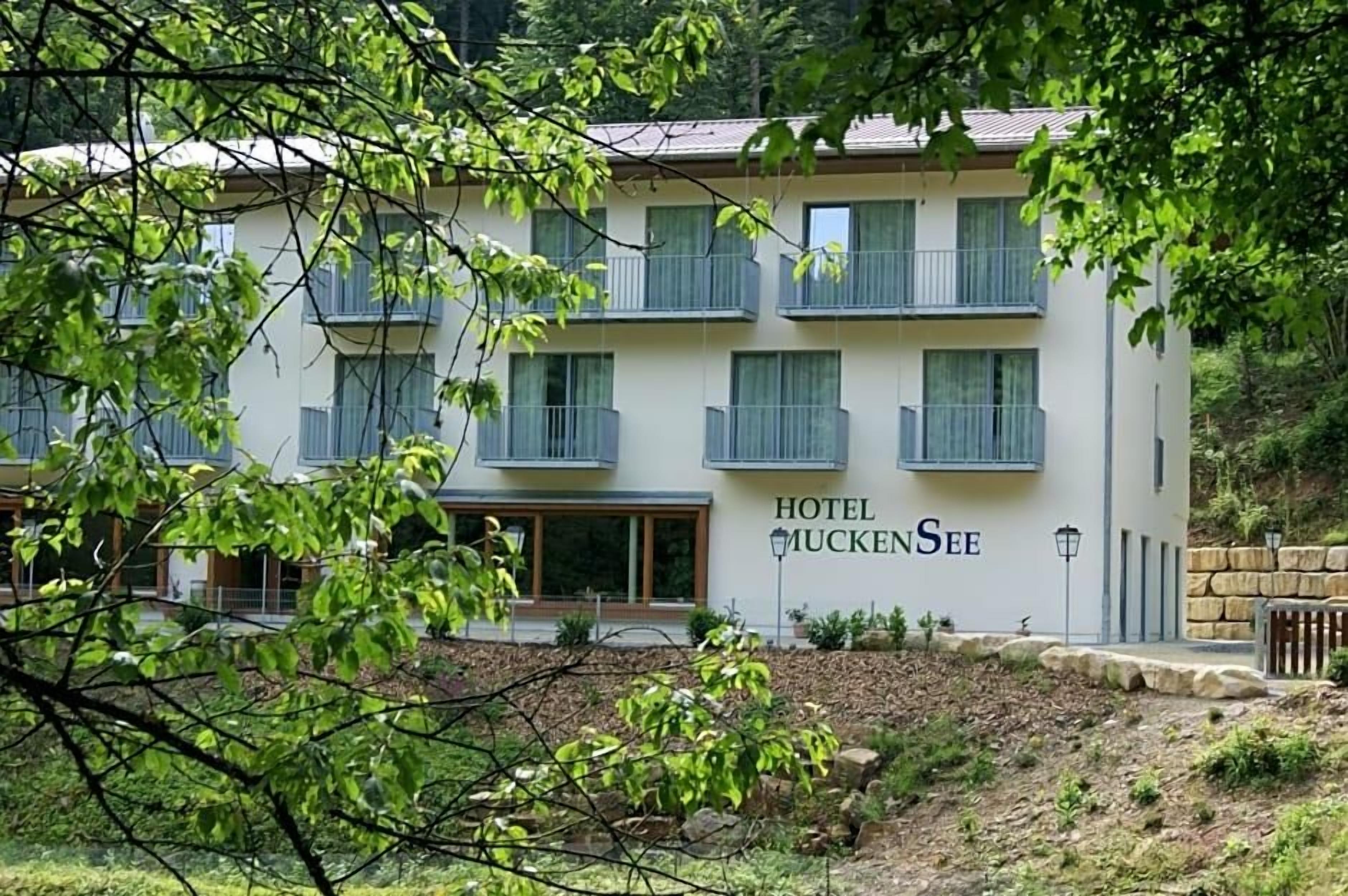 A two-story hotel with balconies, a sign reading 'HOTEL MUCKENSEE,' and a stone wall in the foreground.