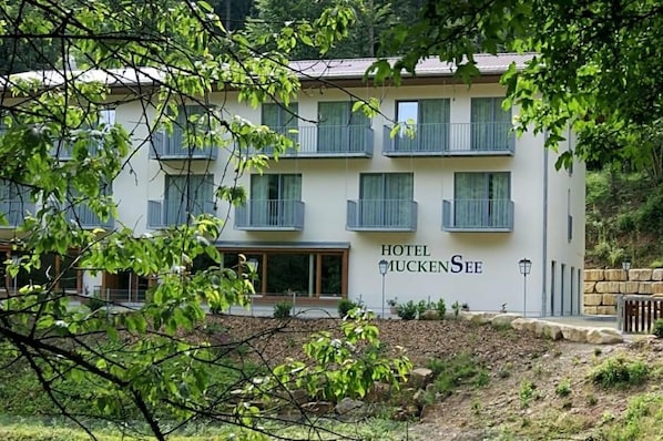 A two-story hotel with balconies, a sign reading 'HOTEL MUCKENSEE,' and a stone wall in the foreground.