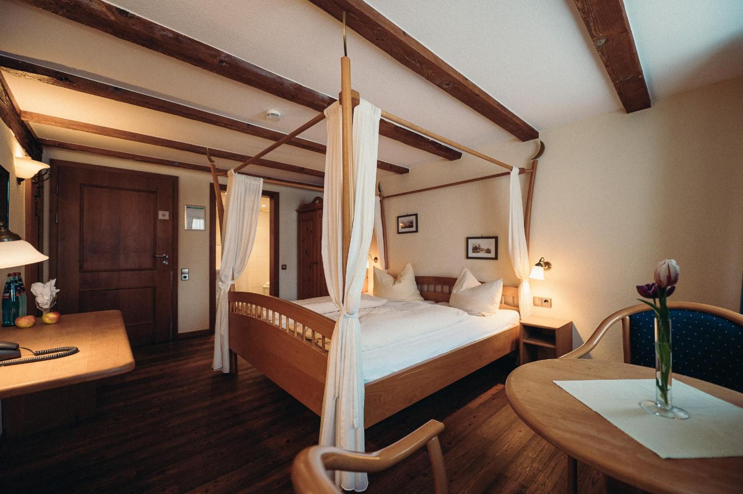 Ein Bett mit Himmelbett, ein Holztisch mit Stuhl, ein Nachttisch und ein kleiner Tisch mit Blumen.