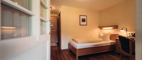A small, cozy bedroom with a single bed, a desk, and a chair. There is a door leading to another room and a window with blinds.