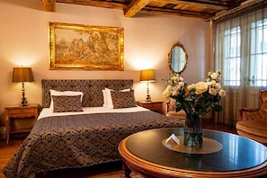 A hotel room with a bed, two bedside tables, a round coffee table, a vase with flowers, a painting on the wall, and a mirror.