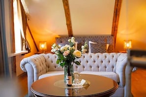 A round table with a vase of flowers and a candle, placed in front of a tufted sofa in a warmly lit room with exposed wooden beams.