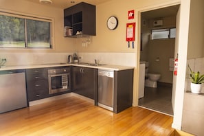 A modern kitchen with a dishwasher, oven, and sink. There is a clock on the wall and a fire blanket hanging on the wall. - Little Norfolk Bay Events And Chalets (Taranna)