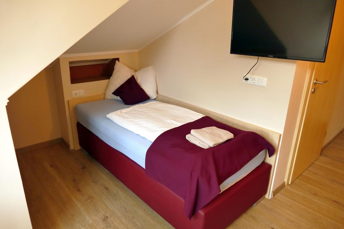 A single bed with a headboard, a flat-screen TV mounted on the wall, and a power outlet on the wall.