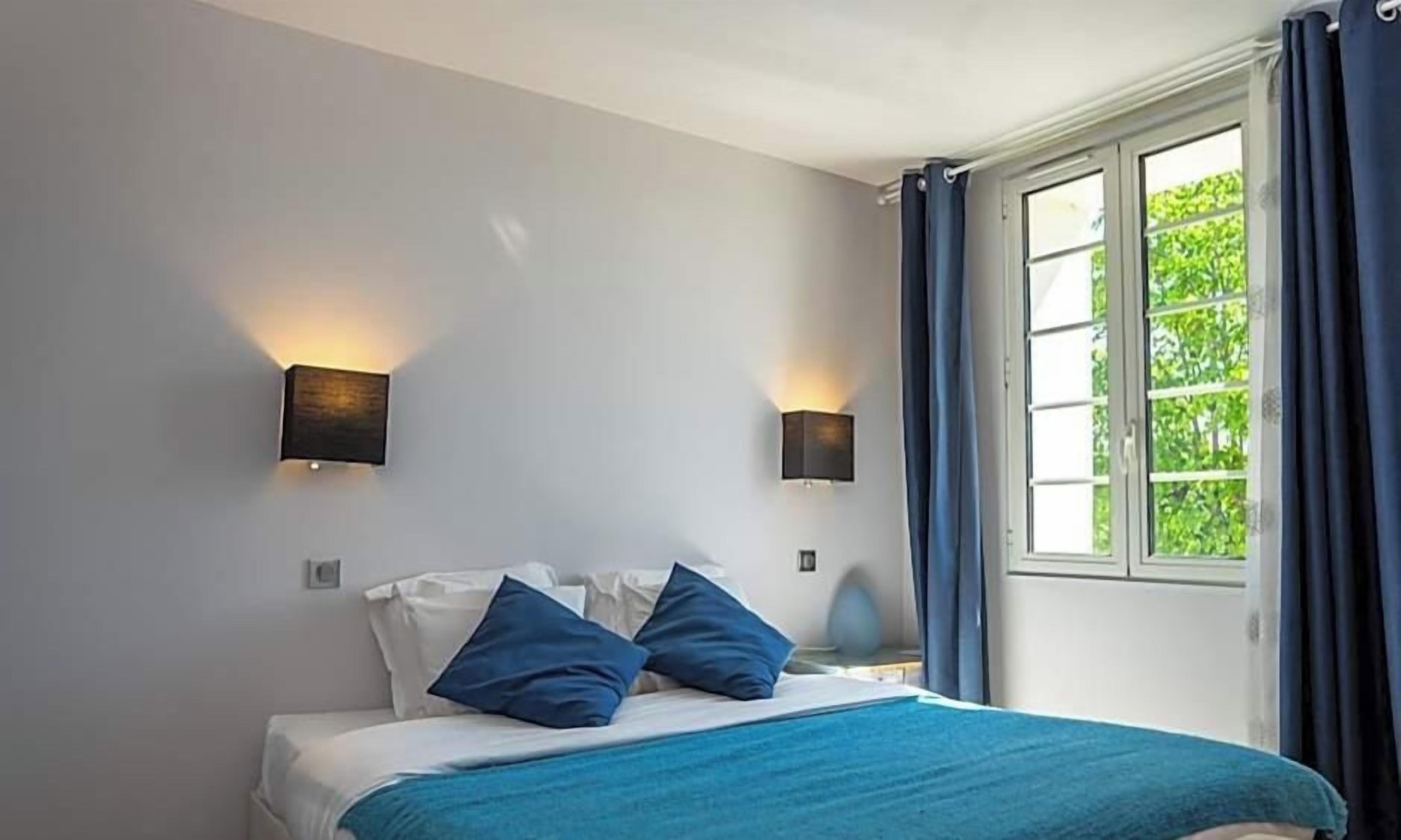 Une chambre à coucher avec un lit, deux lampes fixées au mur, une fenêtre avec des rideaux et une vue sur la verdure à l’extérieur.