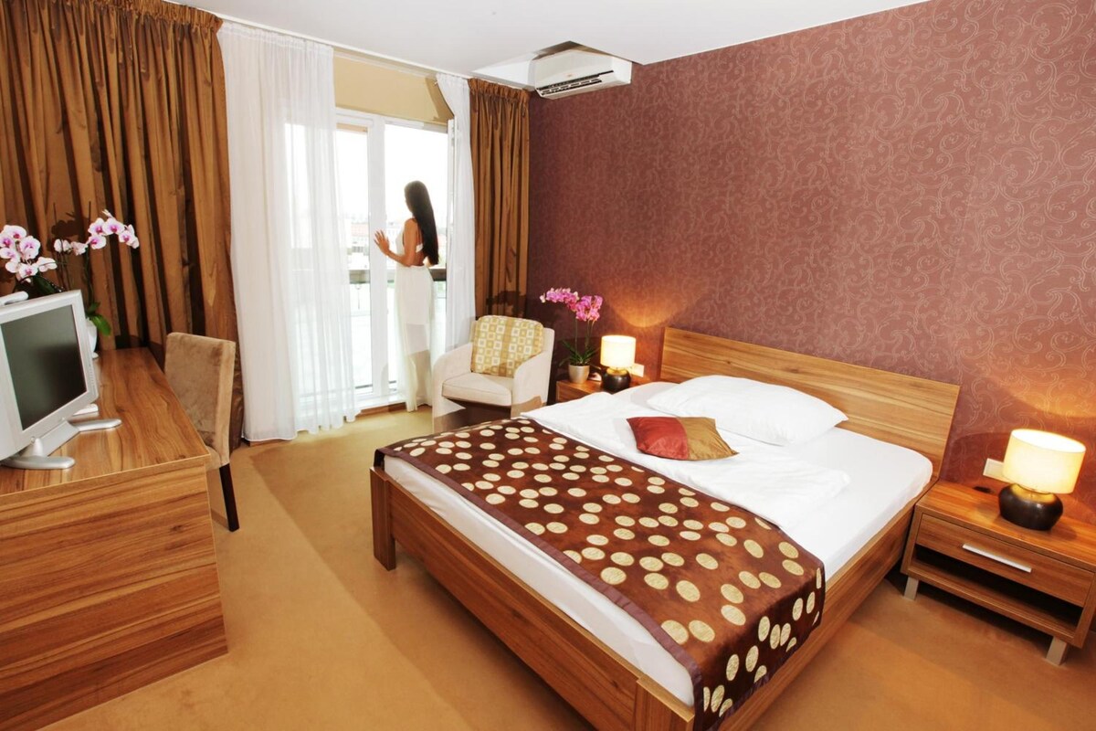 Quarto de hotel com cama, escrivaninha, cadeira, televisão e janela com cortinas.