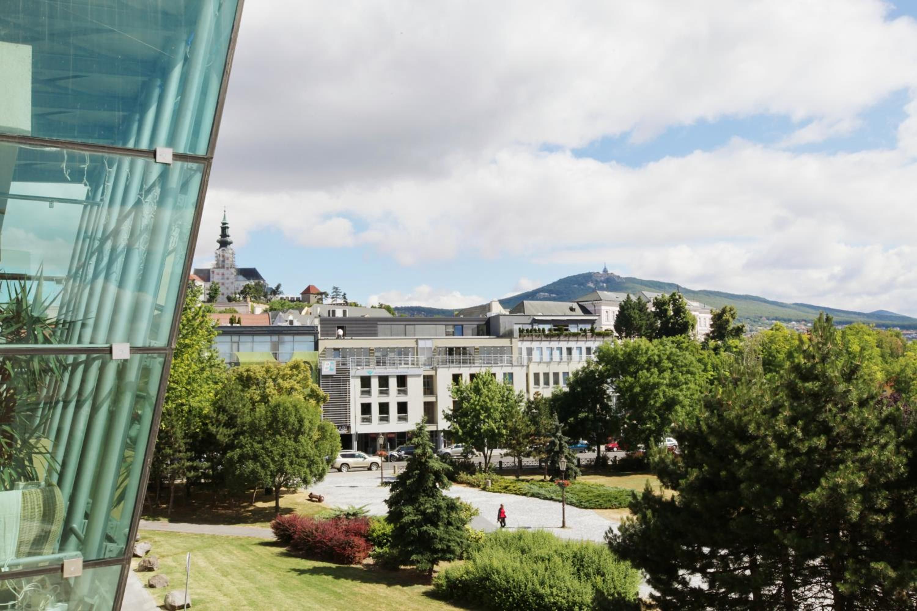 A modern building with a glass facade, a courtyard with trees, and a distant view of a church spire.