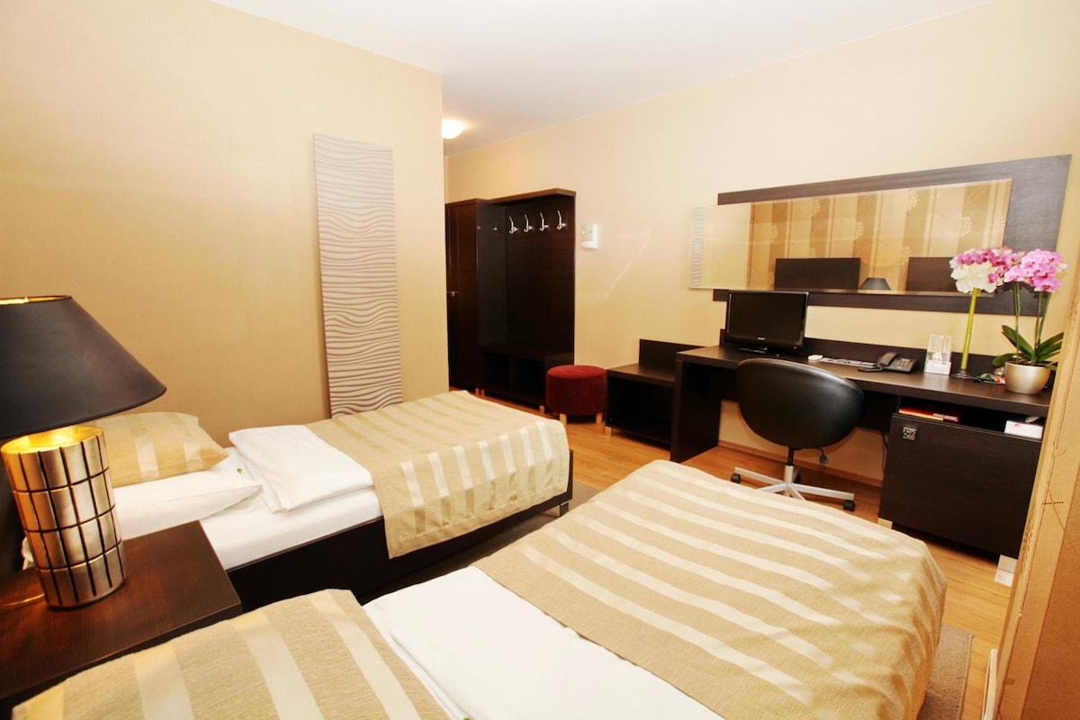 Quarto de hotel com duas camas, uma escrivaninha, uma cadeira, um computador, um abajur e um espelho.