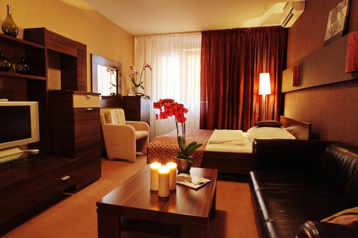 A hotel room with a bed, a sofa, a TV, a desk with a lamp, and a window with curtains.