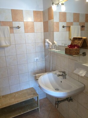 A bathroom with a white sink, a towel rack, a shelf with glassware, and a wicker basket. - Gasthof Pension Golob (Kirchbach)
