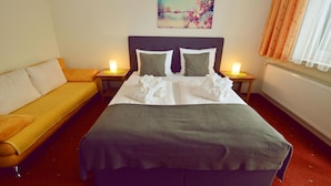 Una habitación de hotel con cama, sofá, dos mesitas de noche con lámparas, un cuadro en la pared y una ventana con cortinas.