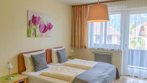Una habitación de hotel con una cama, almohadas, una mesita de noche con lámpara, una ventana con cortinas y un cuadro de flores en la pared.