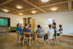 Un grupo de personas sentadas en una sala con techo y paredes de madera, una pantalla de proyector y una pizarra.