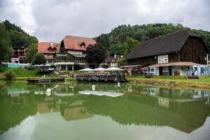 A lakeside view with a wooden building, a restaurant with outdoor seating, and a bridge over the water. - Seegasthof Breineder Familien Seminarhotel (Waldbach-Mönichwald)