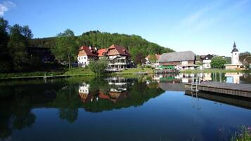 Ein ruhiger See mit Spiegelung von Gebäuden, eine Anlegestelle mit einer Leiter und ein Kirchturm in der Ferne.