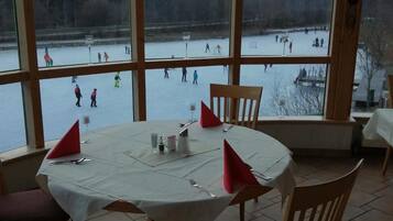 Ein Essbereich mit Tisch für zwei Personen, große Fenster mit Blick auf einen See und Menschen, die Eislaufen.