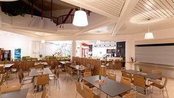 Ein modernes Restaurantambiente mit hohen Decken, Pendelleuchten und verschiedenen Sitzgelegenheiten, darunter Tische und gemütliche Sitzgruppen.