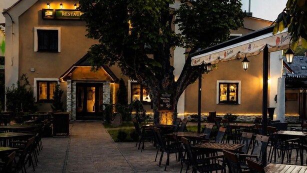 An outdoor restaurant patio with tables and chairs, a building with a 'RESTAURANT' sign, and a tree in the center. - Hotel Restaurant Pod Ka Tany (Krupka)