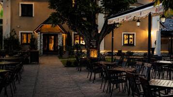 Eine Terrassenfläche eines Restaurants mit Tischen und Stühlen, ein Gebäude mit einem 'RESTAURANT'-Schild und ein Baum in der Mitte.