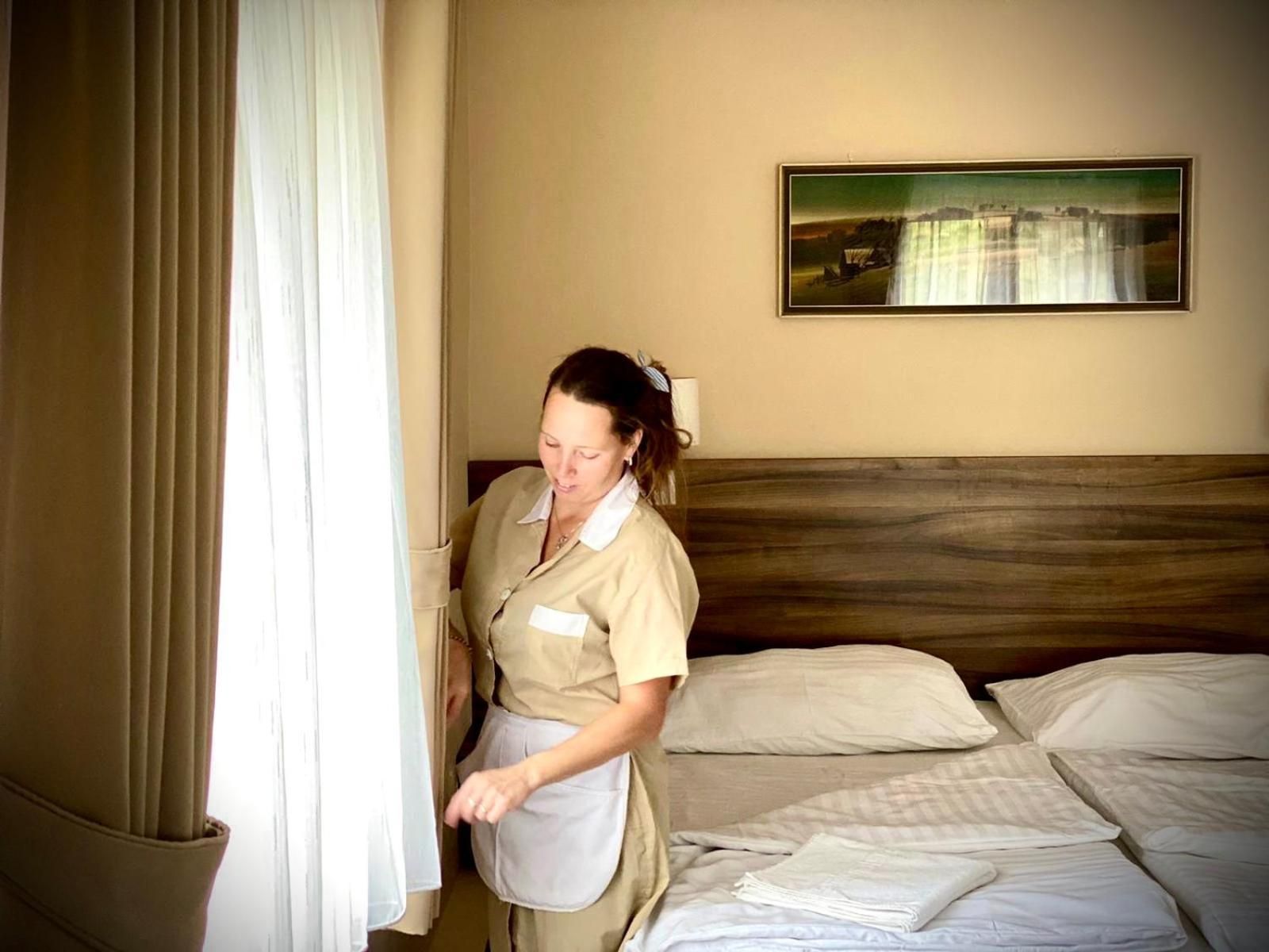 Ein Hotelzimmer mit Bett, Kissen, einem Gemälde an der Wand und einer Person, die am Fenster steht.