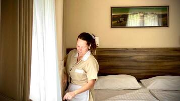 Ein Hotelzimmer mit Bett, Kissen, einem Gemälde an der Wand und einer Person, die am Fenster steht.