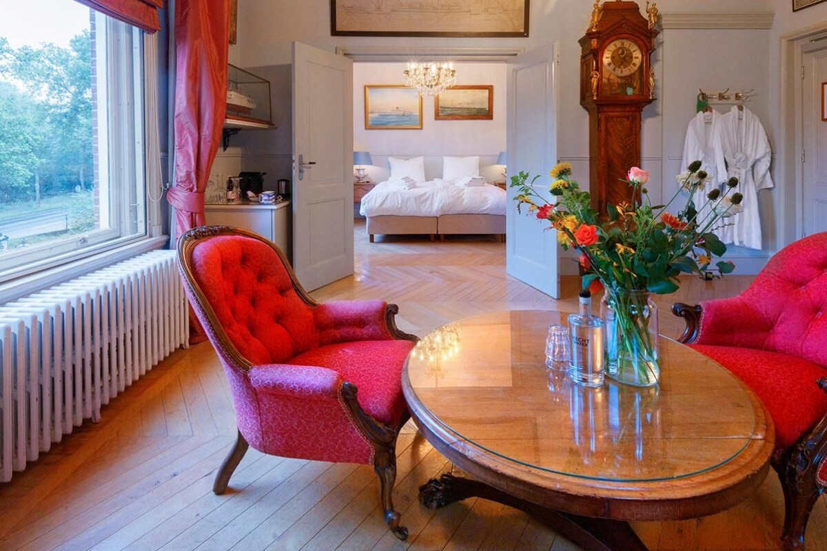 Una habitación con una cama, un ventanal grande, un sillón tapizado en rojo, una mesa redonda de vidrio y piso de madera.